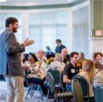 man stands and addresses room of people seated at round tables in Alumni House